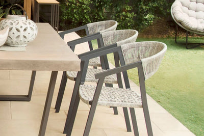 Three outdoor wicker dining chairs with charcoal-coloured aluminium frames are facing an outdoor concrete table with a light on it. The setting is in someone's backyard, with greenery visible in the background.
