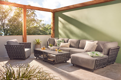 Dark-grey Lawson 7-seater outdoor rope modular lounge with a mix of beige, grey, and green throw pillows, surrounding two Lawson outdoor coffee tables set with tea, a pot plant, and fruit. The lounge is set in a sunny outdoor area of a house with a green-painted wall, and trees peek through, creating a peaceful and inviting outdoor living space.