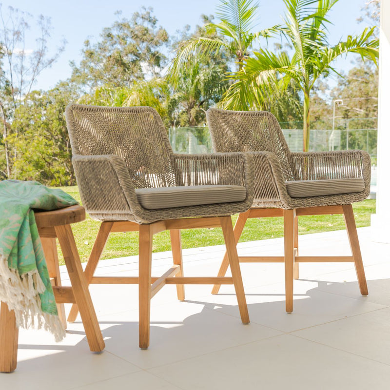 A couple of wicker dining chairs with natural frame, light grey fabric and white wicker set outdoors with trees in the background. Outdoor furniture, outdoor dining furniture, wicker chair.