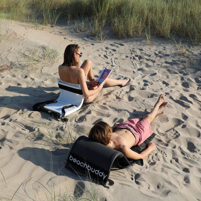 A mother and son relaxing on the beach using outdoor backrests. The mother is on a white 'Beach Buddy' backrest (which functions as a portable sun lounger), while the son uses a black one. Both are seated on the sand, enjoying the sunny weather and relaxing together.