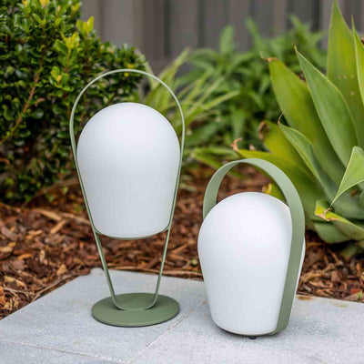 2 chic aluminium 'Bobby' outdoor lamps in green, one mid-high and one low, set on stone tiling in an outdoor area with lush greenery in the background.