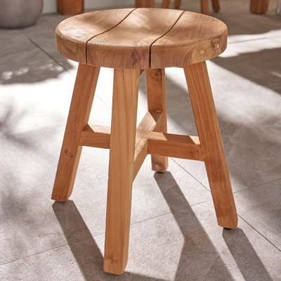 A round teak stool set in a sunny outdoor area, with the natural wood glowing under the sunlight, showcasing the elegance of teak wood furniture in a relaxed outdoor space.