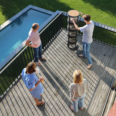 5-piece 'Casper' balcony furniture collection stacked in a single pile, featuring four stackable stools with soft cushioning and a matching coffee table with a wooden teak top. A family of four is either unpacking or packing up the furniture, standing around it on an outdoor wooden deck.