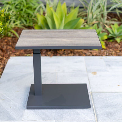 Black and grey 60 cm aluminium 'Clifton' pop-up outdoor side table with a ceramic tabletop, set in a backyard area with plants in the background.
