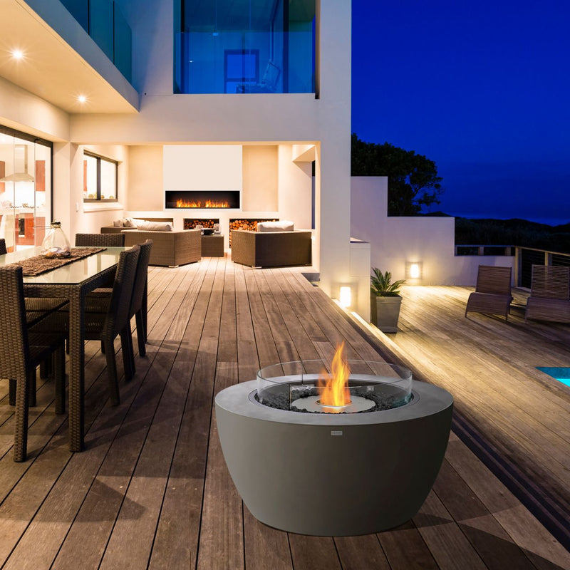 Cylindrical grey EcoSmart Pod 40 firepit set in the outdoor area of a modern house by an outdoor dining table, adding warmth to the outdoor furniture layout.