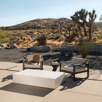 A 165 cm rectangular outdoor concrete coffee table in white, part of our 'Blinde' range, is set in an outdoor desert area. The table is accompanied by two outdoor aluminium chairs and a small side table with two books and a plant on it. In the background, the desert landscape features sand, dry plants, and cacti. The furniture is viewed from a side angle.