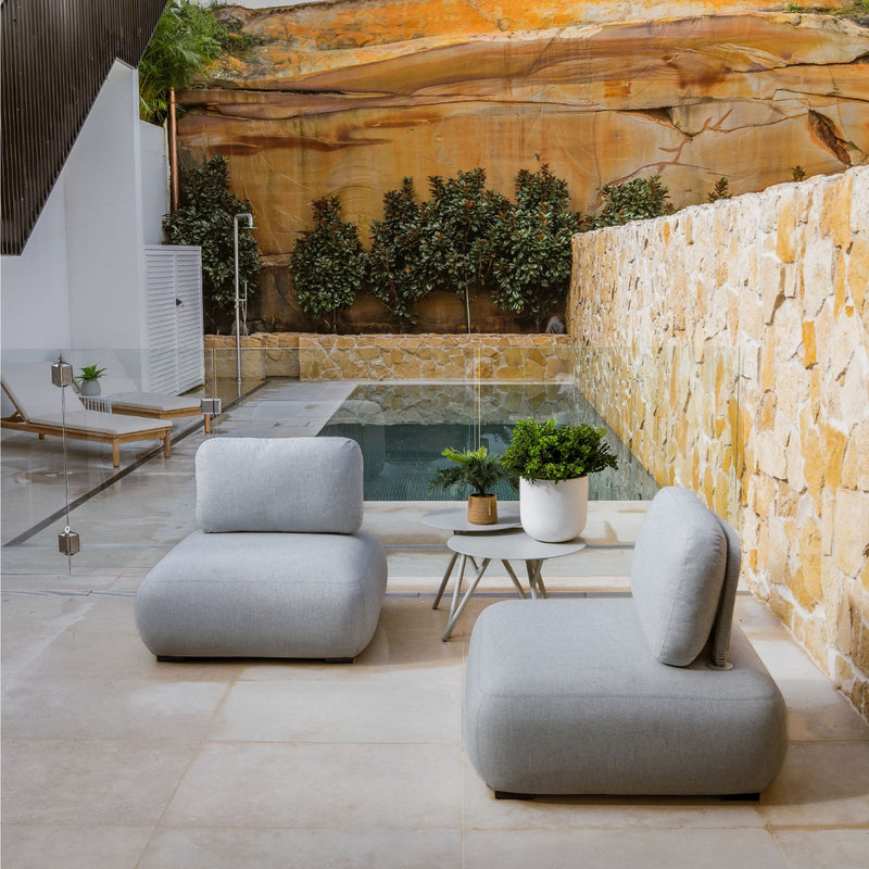 Two grey Iowa Outdoor Upholstered Lounge Chairs accompanied by two grey Iowa Outdoor Aluminium Round Coffee Tables, all set poolside in a modern European home with sandstone walls. The orange hues of the furniture contrast beautifully with the greys and blues of the water, creating an inviting and stylish outdoor living space.