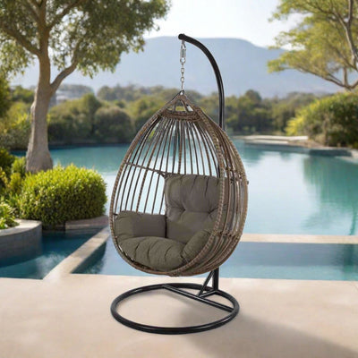 A cozy outdoor scene featuring a wicker hanging egg chair with plush gray cushions, suspended from a sturdy stand. The chair is placed in an outdoor setting with green foliage in the background, exuding a tranquil vibe.