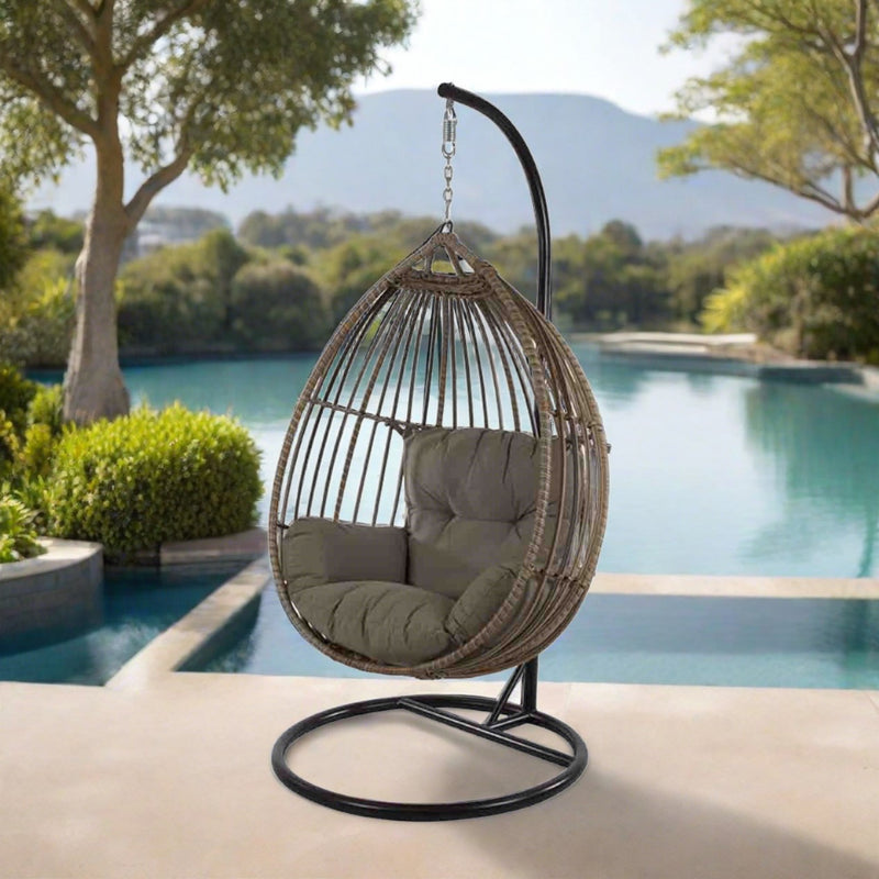 A cozy outdoor scene featuring a wicker hanging egg chair with plush gray cushions, suspended from a sturdy stand. The chair is placed in an outdoor setting with green foliage in the background, exuding a tranquil vibe.