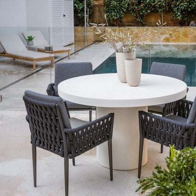 Four dark grey Lawson outdoor rope dining chairs around a white round concrete table with two pot plants, set poolside, highlighting a stylish alfresco furniture setup.