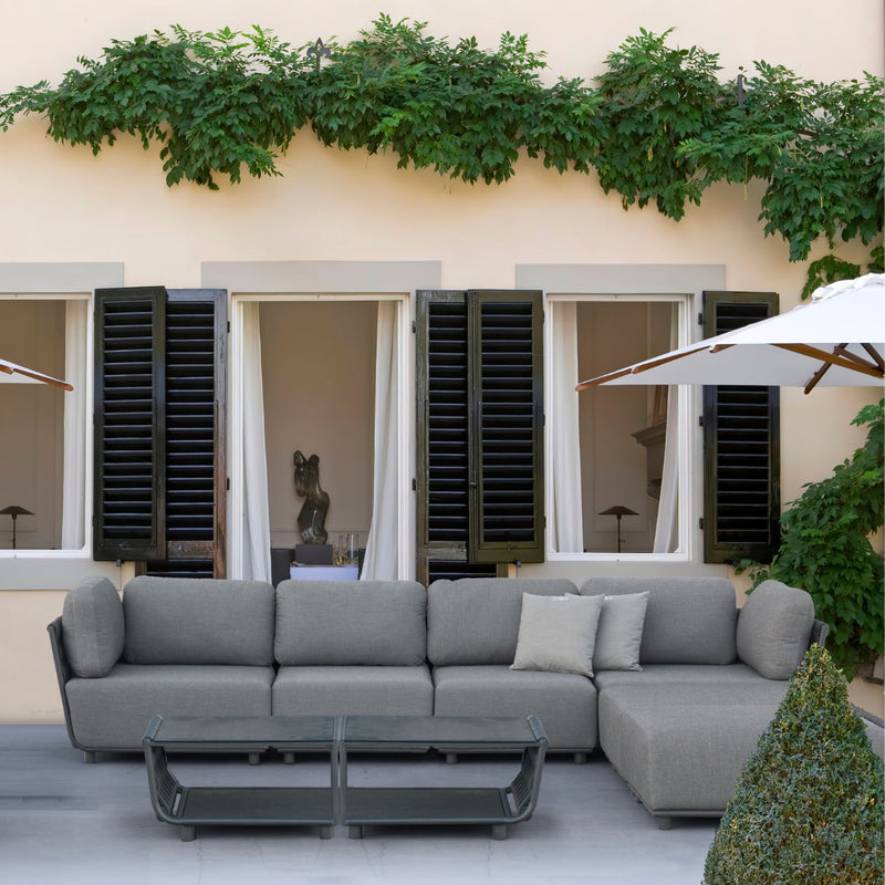 Dark-grey Lawson 7-seater outdoor rope modular lounge with two grey throw pillows, accompanied by two Lawson outdoor coffee tables, all set in an outdoor living space protected by an umbrella. The background features a house adorned with greenery, creating a serene and stylish alfresco environment.