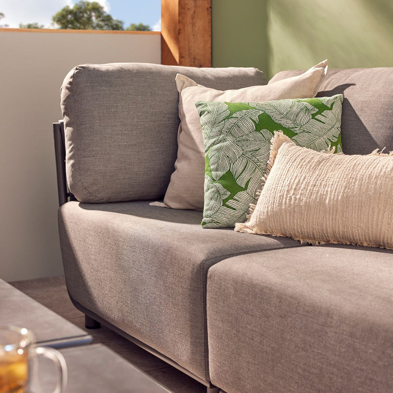 Close-up of a dark grey Lawson 4-seater outdoor rope modular lounge, decorated with three throw pillows, set in an outdoor living space with the sun shining in. The lounge is placed against a green wall, with a glimpse of a coffee table in the bottom left corner, featuring a tea set, creating a cozy and stylish alfresco setting.