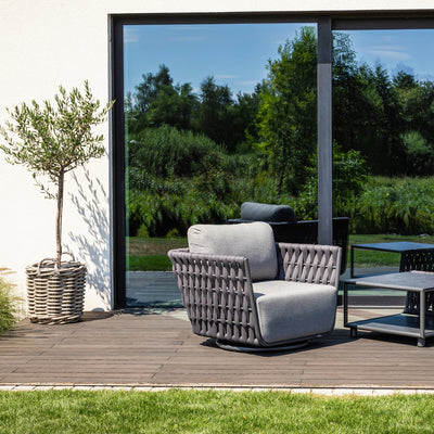 Stylish Lawson Outdoor Rope Armchair in charcoal, featuring a woven rope design and soft gray cushions, placed on a wooden deck next to a wicker pot with a small olive tree. The background includes large glass doors reflecting lush greenery.