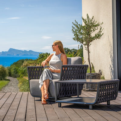 Lawson Outdoor Rope Armchair in charcoal, with a woven rope design and soft gray cushions, paired with the matching Lawson Outdoor Rope Coffee Table. A woman relaxes on the armchair, enjoying a stunning ocean view, surrounded by greenery and a potted olive tree on a wooden deck.