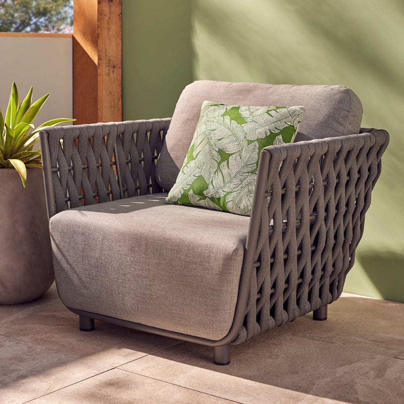 Outdoor furniture from the Lawson Collection featuring a rope outdoor lounge chair, armchair, ottoman, and coffee table, all in a backyard setting with a chair with a pillow and a potted plant.