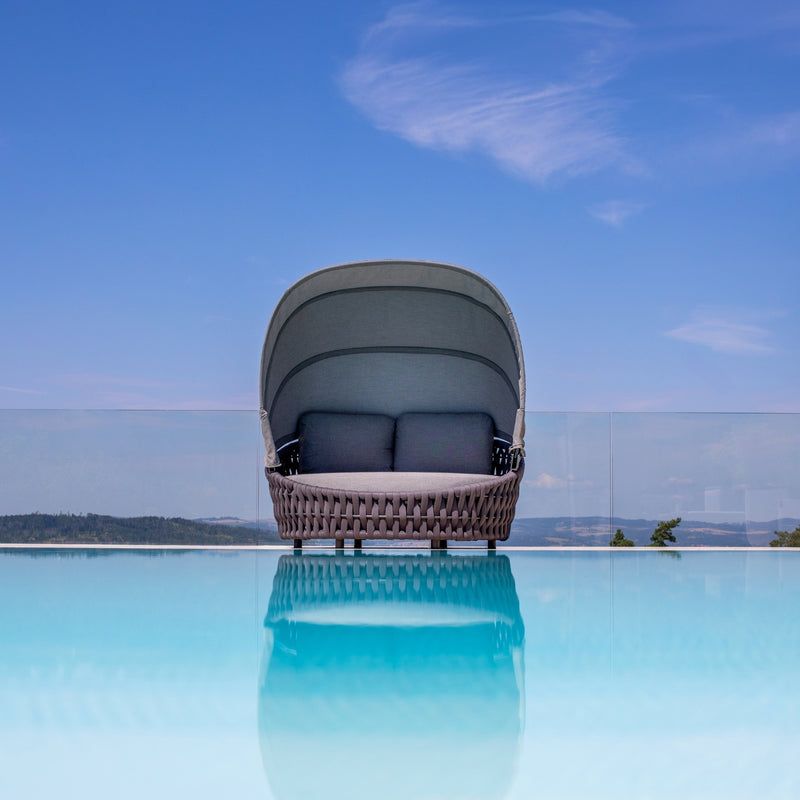 Rope daybed with a curved canopy, two gray cushions, set against a serene blue infinity pool reflecting the sky, overlooking distant hills, embodying the essence of luxurious outdoor relaxation.