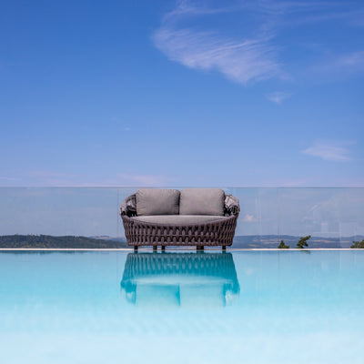 
Rope daybed with two gray cushions, set against a glass railing and a pristine infinity pool reflecting the clear blue sky, offering a seamless view of the distant rolling hills and serene outdoor ambiance.