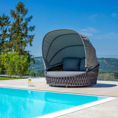 Rope daybed with a curved canopy and gray cushions, positioned by a sparkling blue pool, surrounded by lush greenery and scenic hills, evoking a sense of outdoor comfort and tranquility.