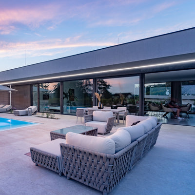 Taupe outdoor lounge set with light sand-coloured cushions and rope detailing, featuring a 4-seater sofa, matching armchairs, and coffee table, arranged on a spacious tiled patio beside a glowing pool, with modern architecture and large glass doors reflecting the vibrant hues of a sunset sky.
