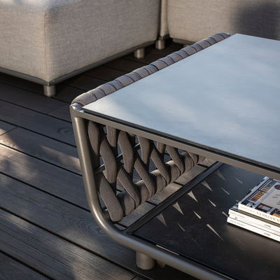Close-up of a modern outdoor coffee table featuring charcoal rope detailing, a sleek grey tabletop, and a lower shelf, placed on a dark timber deck alongside a grey sectional sofa, showcasing elegant craftsmanship and functionality.