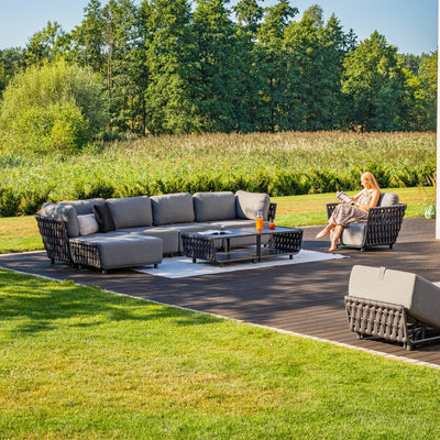 Spacious outdoor 7-seater sectional sofa with charcoal rope detailing and grey cushions, arranged on a wooden deck surrounded by lush greenery, complemented by a matching coffee table, creating a sleek and modern outdoor lounging area.