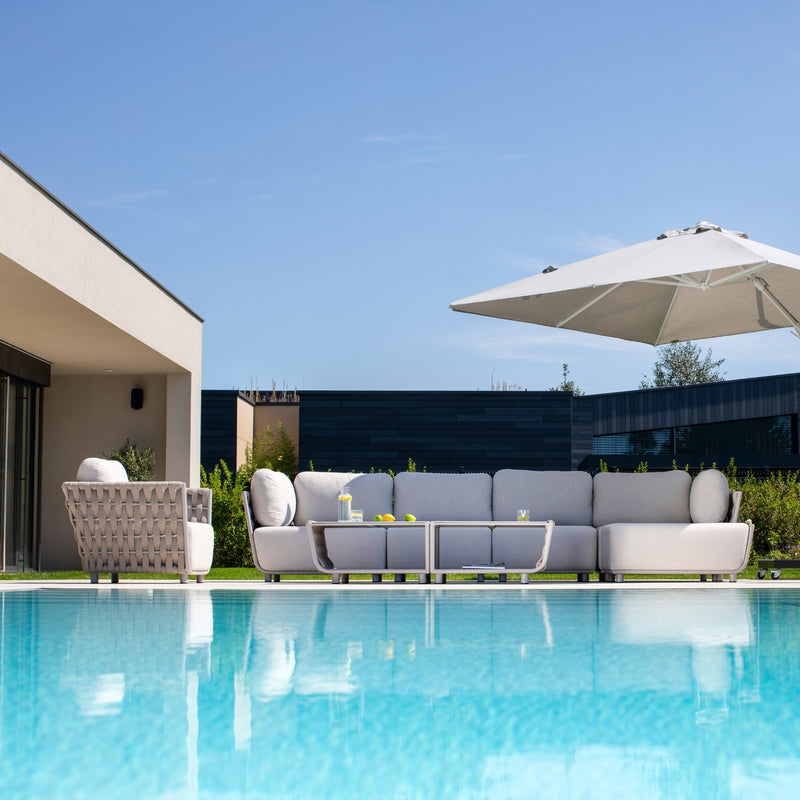 Outdoor modular sofa set in taupe with light sand-coloured cushions and matching rope-detailed armchair, paired with a sleek coffee table, set on a poolside patio under a large white umbrella, surrounded by lush greenery and modern architecture under a clear blue sky.