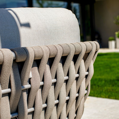 Close-up of a taupe rope weave on the backrest of an outdoor lounge chair, paired with light sand-coloured cushions, set against a backdrop of lush green grass and a modern patio, showcasing intricate craftsmanship and contemporary design.