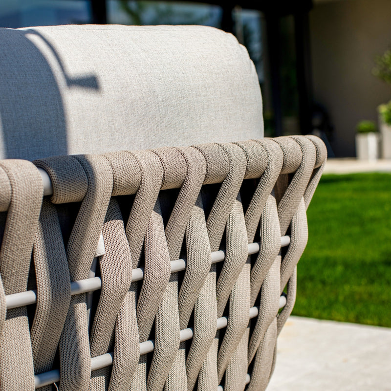 Close-up of a taupe rope weave on the backrest of an outdoor lounge chair, paired with light sand-coloured cushions, set against a backdrop of lush green grass and a modern patio, showcasing intricate craftsmanship and contemporary design.