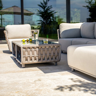Close-up of an outdoor coffee table with taupe rope detailing and a glass top, adorned with fresh limes, a glass pitcher of water, and a drink glass, surrounded by a taupe modular sofa and armchair with light sand-coloured cushions, set on a tiled patio reflecting greenery and modern architecture for a stylish outdoor setting.