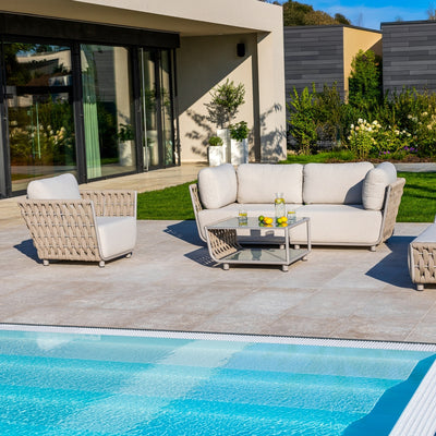Taupe outdoor 2-seater sofa with light sand-coloured cushions and rope detailing, placed on a tiled patio near a pool, complemented by a matching coffee table and vibrant greenery, creating a luxurious and inviting outdoor living space.