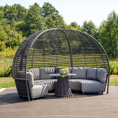 Outdoor rope pavilion lounge featuring a dome-shaped structure with a woven rope design, gray cushions, and a circular woven table holding a bowl of lemons, set on a wooden deck surrounded by lush greenery under a bright blue sky.