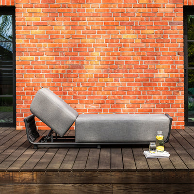 Outdoor grey sun lounger with an adjustable backrest, featuring sleek rope accents, placed on a wooden deck against a vibrant red brick wall. A side setup includes a tray with a pitcher of lemon water, a glass, and a magazine, evoking a stylish and tranquil relaxation scene.