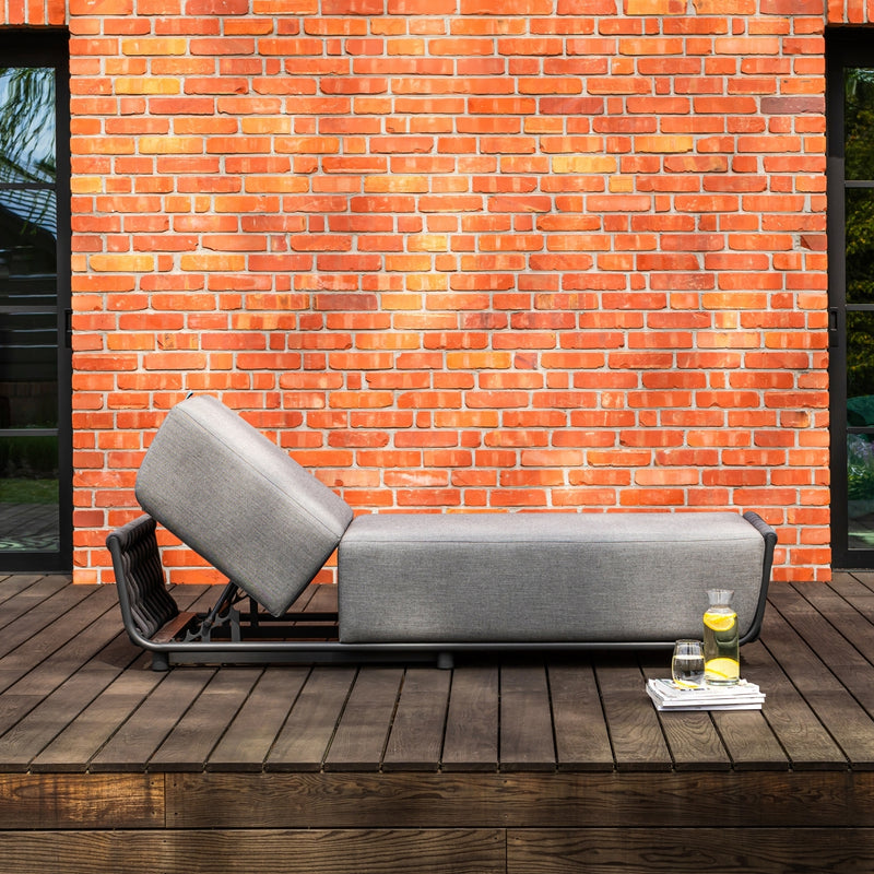 Outdoor grey sun lounger with an adjustable backrest, featuring sleek rope accents, placed on a wooden deck against a vibrant red brick wall. A side setup includes a tray with a pitcher of lemon water, a glass, and a magazine, evoking a stylish and tranquil relaxation scene.