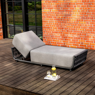 Outdoor grey sun lounger with an adjustable backrest and rope detailing, set on a timber deck against a red brick wall with large, black-framed windows, accompanied by a pitcher of lemon water, a glass, and magazines for a stylish outdoor retreat.