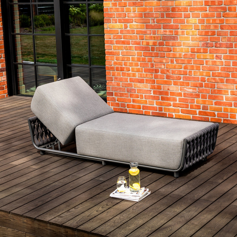Outdoor grey sun lounger with an adjustable backrest and rope detailing, set on a timber deck against a red brick wall with large, black-framed windows, accompanied by a pitcher of lemon water, a glass, and magazines for a stylish outdoor retreat.