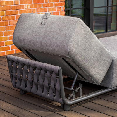 Close-up of an adjustable grey sun lounger with intricate rope detailing and a sturdy metal frame, positioned on a timber deck against a vibrant red brick wall and black-framed glass windows, showcasing functional design and outdoor sophistication.