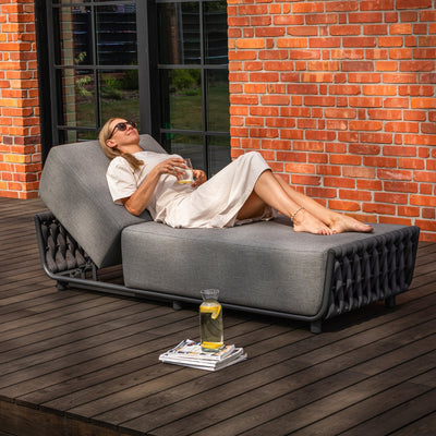 A woman relaxing on a grey sun lounger with an adjustable backrest and rope accents, holding a drink, placed on a timber deck against a vibrant red brick wall with black-framed glass windows, exuding comfort and outdoor elegance.