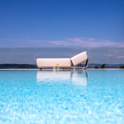 Taupe outdoor sun lounger with light sand-coloured cushions and an adjustable backrest, positioned by a pool with a glass balustrade, overlooking a serene landscape under a vibrant blue sky, embodying luxurious relaxation.