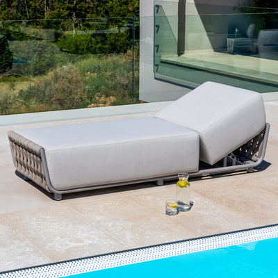 Taupe outdoor sun lounger with light sand-coloured cushions and elegant rope detailing, positioned on a tiled patio beside a shimmering pool, with a glass balustrade and lush greenery in the background, exuding modern outdoor sophistication.