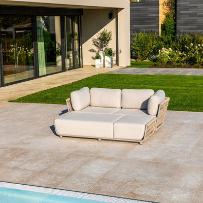 Outdoor Taupe sofa with rope detailing and plush light sand cushions, set on a tiled patio by a pool, surrounded by lush greenery, a neatly manicured lawn, and modern architecture featuring large glass doors and sleek design elements.