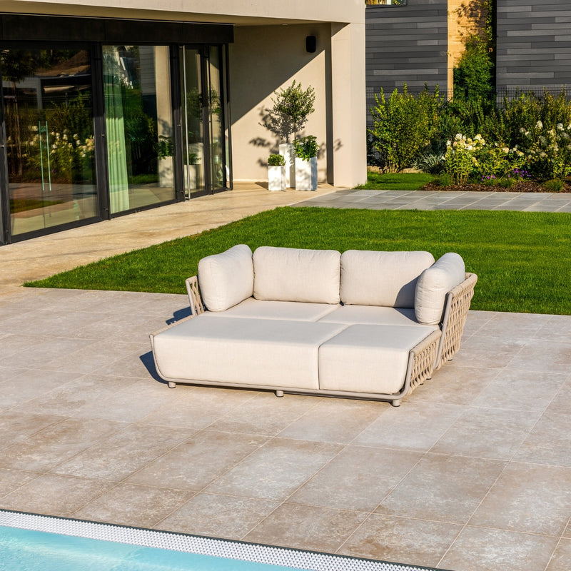 Outdoor Taupe sofa with rope detailing and plush light sand cushions, set on a tiled patio by a pool, surrounded by lush greenery, a neatly manicured lawn, and modern architecture featuring large glass doors and sleek design elements.