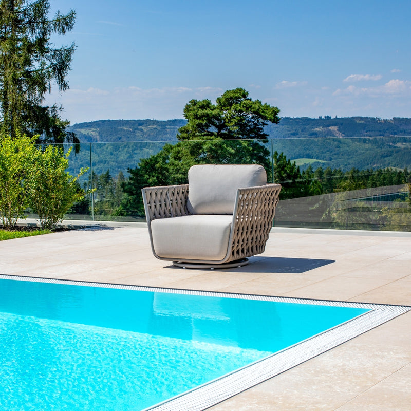 Single outdoor rope swivel chair in Taupe placed beside a shimmering blue pool, set against a backdrop of lush green hills, clear glass fencing, and a bright summer sky, embodying relaxation and outdoor elegance.