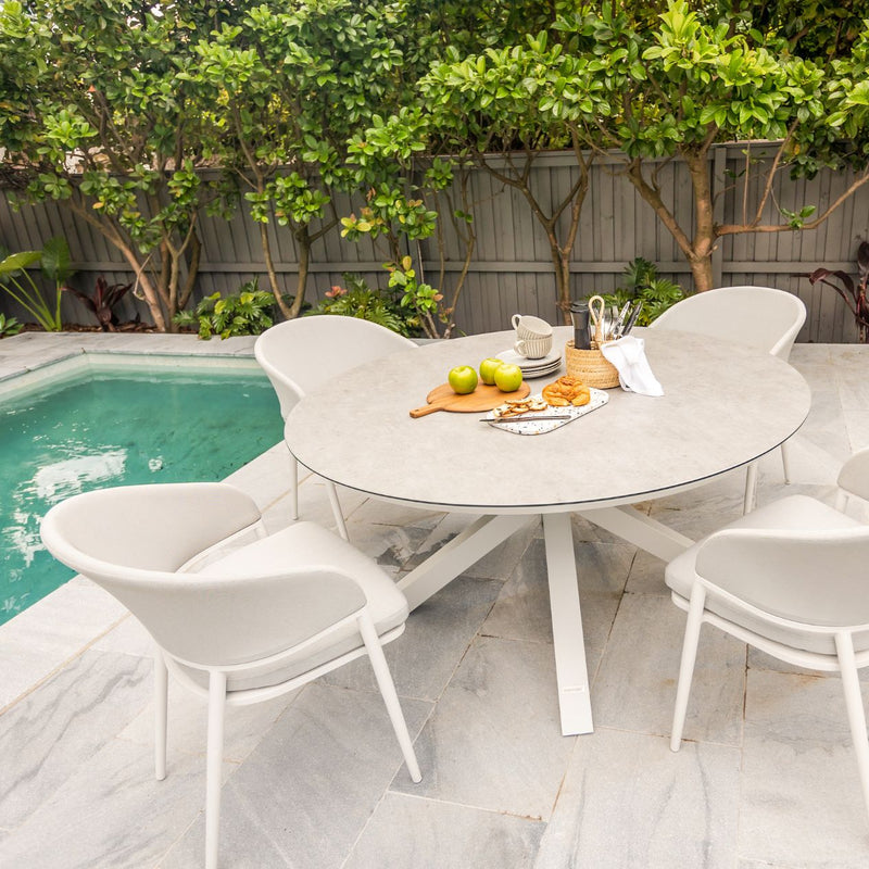 Four white aluminum Medea outdoor dining chairs with sleek zip-off cushion covers surrounding an outdoor dining table, making a complete outdoor dining set. The table is set with food and cutlery, positioned by a pool with greenery and trees in the background.