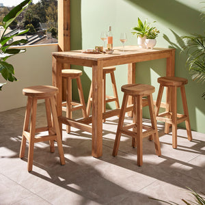 The 150 cm 'Miles' outdoor bar table, a favourite among Remarkable Outdoor Living's wooden outdoor tables, is shown with five wooden stools arranged around it. This setup is positioned against a green wall in an outdoor area. The scene captures the table with wine and cheese, bathed in sunlight, with lush greenery surrounding the area.