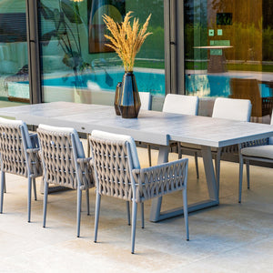 The Mykonos Outdoor Aluminium Extension Table in light gray is paired with Eight woven armchairs featuring beige braided details and gray cushions. Set on a tiled patio, it’s decorated with vases of pampas grass and overlooks a lush lawn and landscaped garden.