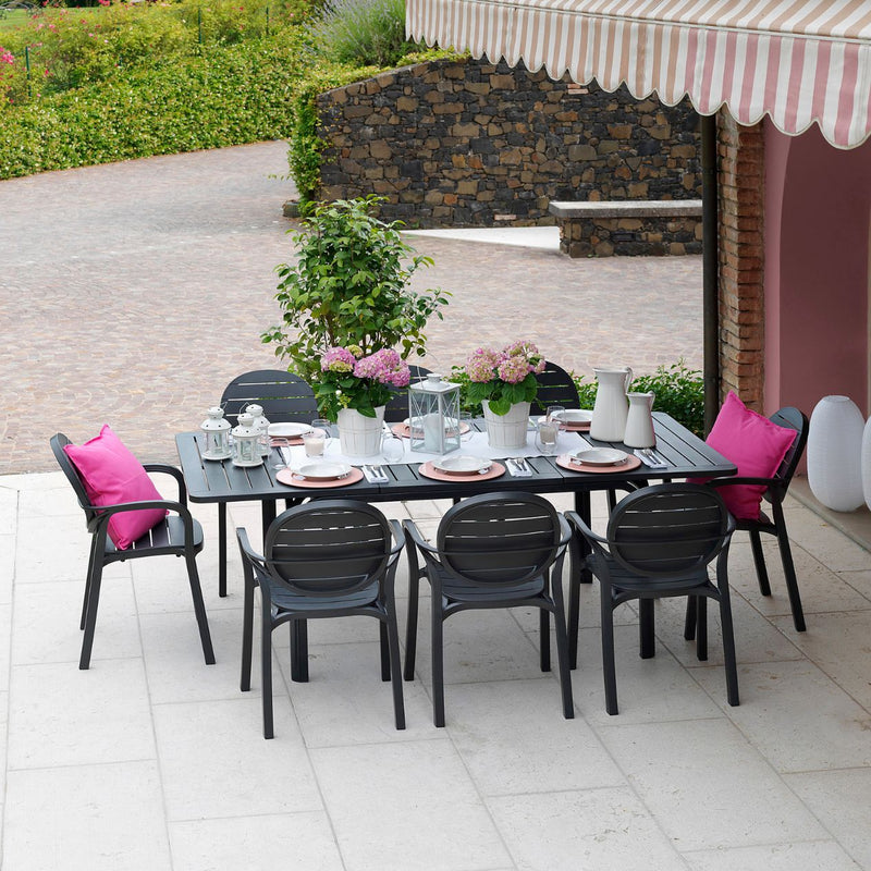 Black 210/280 cm Nardi Alloro Resin Extension Dining Table with eight matching chairs and pink pillows, set on a concrete outdoor area surrounded by greenery, styled with cutlery and flowers, perfect for alfresco furniture setups.