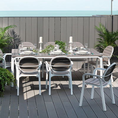 Elegant white and brown Nardi Alloro Resin Extension Dining Table, perfect for alfresco furniture gatherings, with eight matching chairs on a deck with greenery and a scenic seaside backdrop, table set with cutlery and flowers.