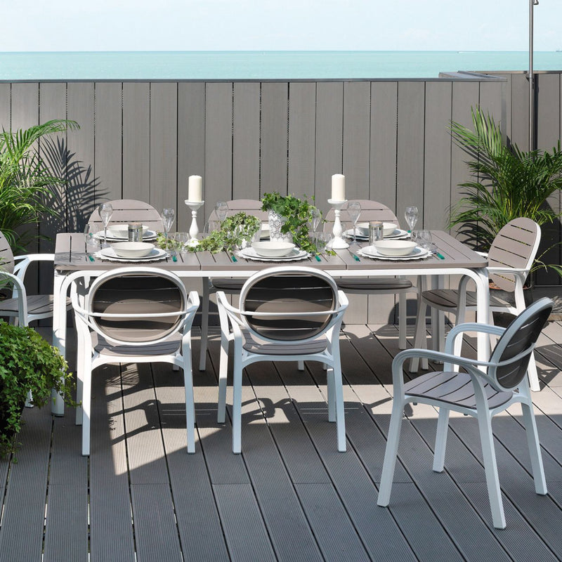 Elegant white and brown Nardi Alloro Resin Extension Dining Table, perfect for alfresco furniture gatherings, with eight matching chairs on a deck with greenery and a scenic seaside backdrop, table set with cutlery and flowers.