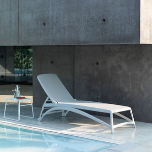 White resin 'Nardi' sun lounger with a textilene seat and backrest, accompanied by a matching coffee table with fruit and water on it, set poolside in a modern house.