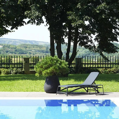 A single black resin 'Nardi' sun lounger with a textilene seat and backrest, accompanied by a matching coffee table, set poolside in an outdoor area with greenery in the background.